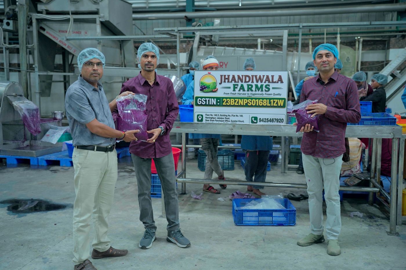 The Jhandiwala Farms team on the processing floor holding sealed packs beside the company signboard, with the wash line and crates behind them.